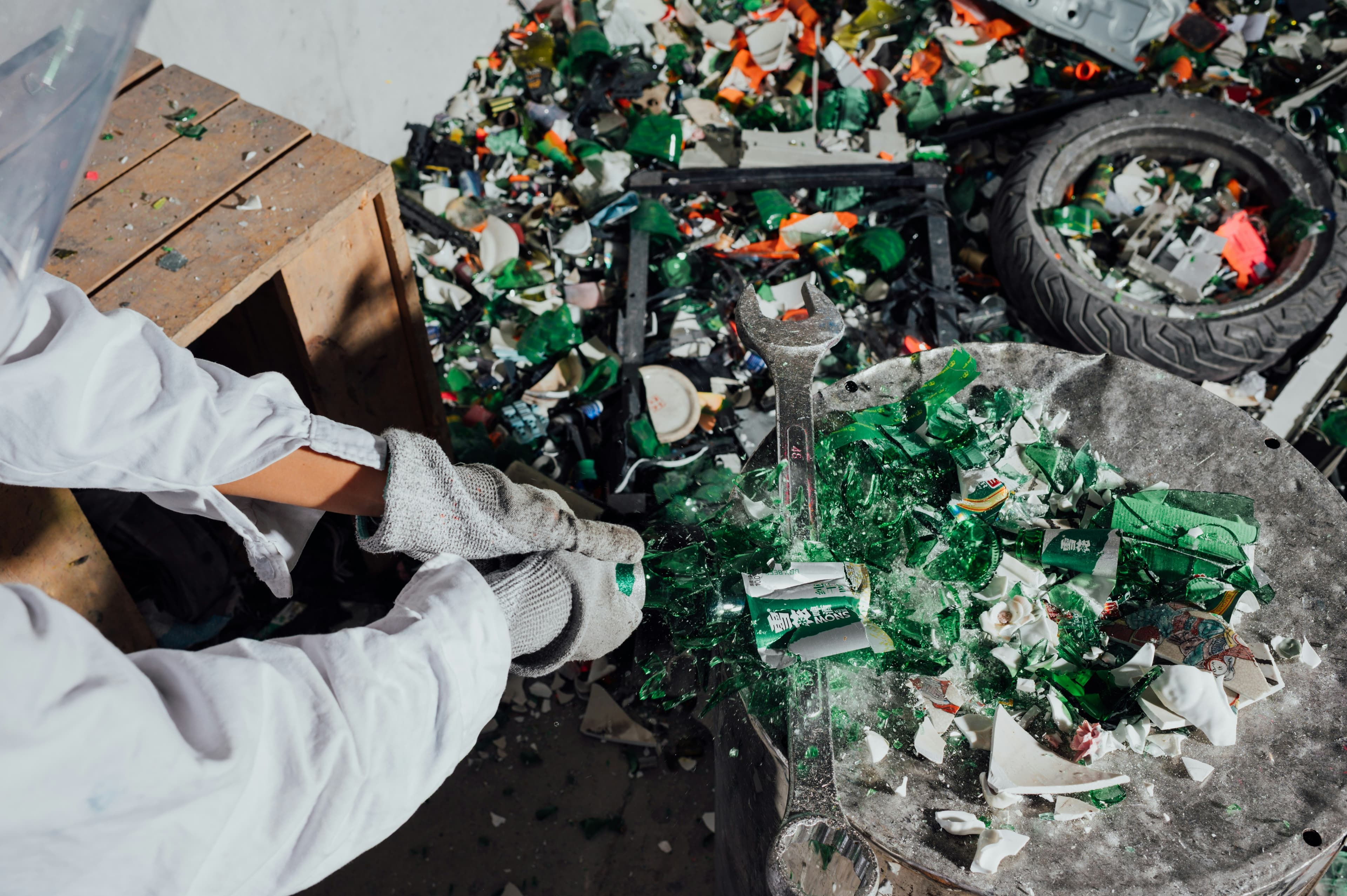 A gloved worker breaking green glass bottles on a metal drum beside piles of sorted scrap