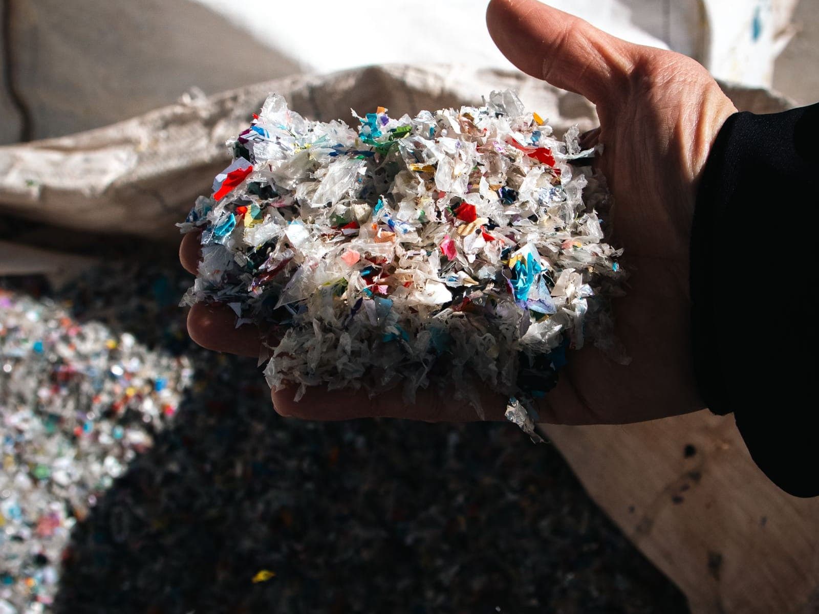 A hand holding shredded, multicoloured plastic flakes above an open bulk bag of sorted plastic