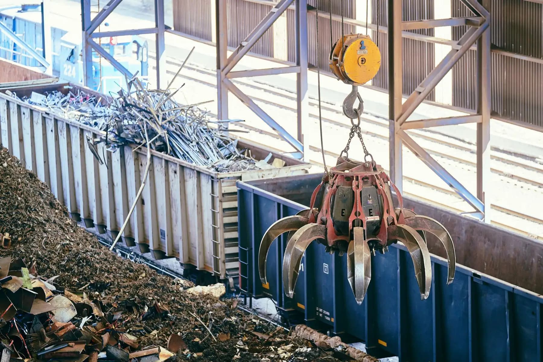 An overhead crane grab lifting scrap metal into a wagon inside a recycling shed